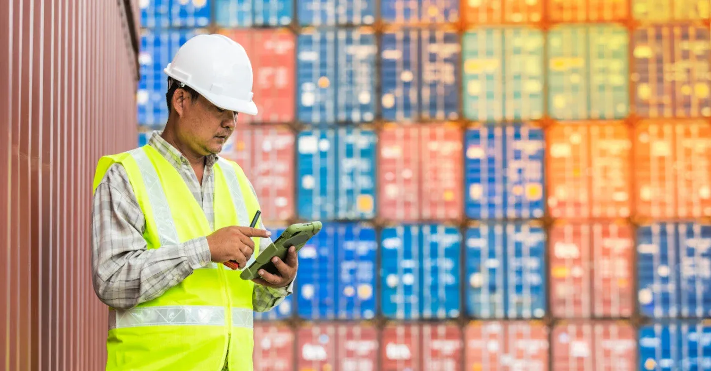 man in hard hat and construction vest taking notes in front of cargo containers