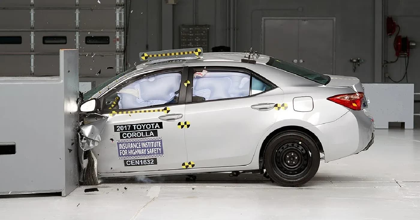 A silver 2017 Toyota Corolla undergoing a crash test, colliding head-on with a solid barrier. The front of the car is crumpled, side airbags are deployed, and the car bears labels from the Insurance Institute for Highway Safety. The scene takes place in a controlled indoor crash testing facility.